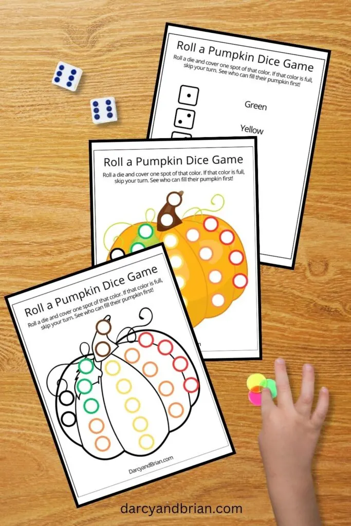 Digital mockup image of the two different pumpkin game mats and the dice legend overlapping each other on a table. Pair of dice, a few bingo chips, and a child's hand are placed near the pages.