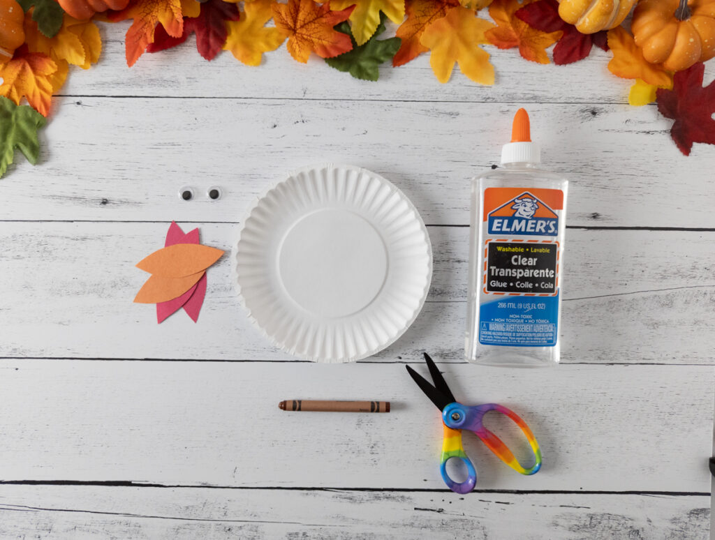 Supplies for making the rocking turkey: construction paper feathers, googly eyes, small paper plate, brown crayon, scissors, and a bottle of glue.