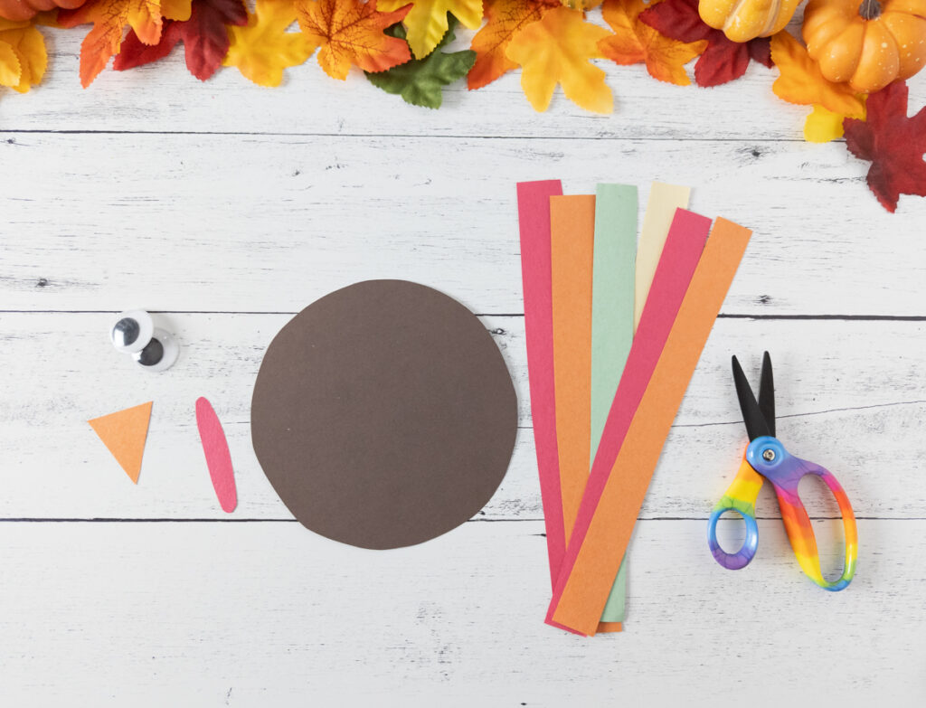 Brown circle in the middle. Strips of red, orange, yellow, and green paper on the right with scissors. On the left are googly eyes, a small orange triangle and a skinny red oval cut out.