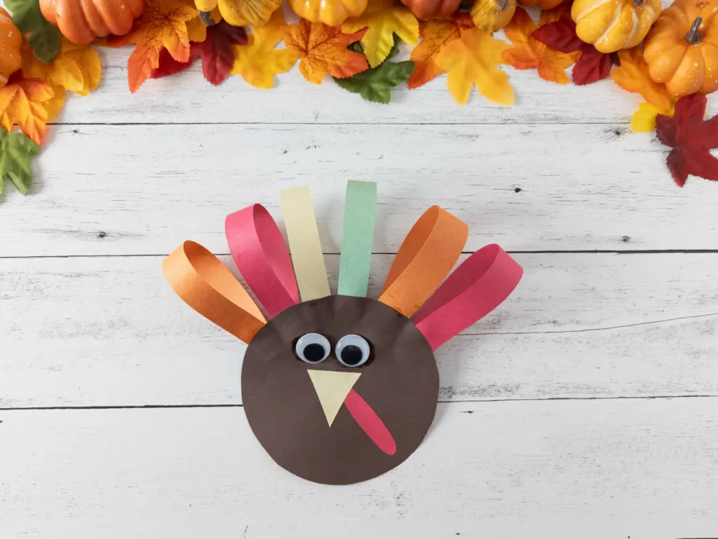 Completed preschool paper turkey craft. Colorful faux leaves and pumpkins decorate the top edge of the photo.