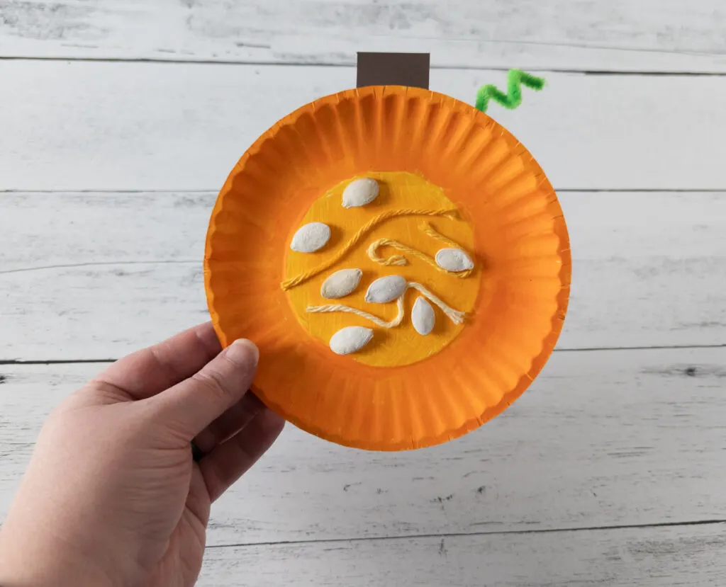 White woman's hand holding up finished pumpkin craft made using a small paper plate and pumpkin seeds.