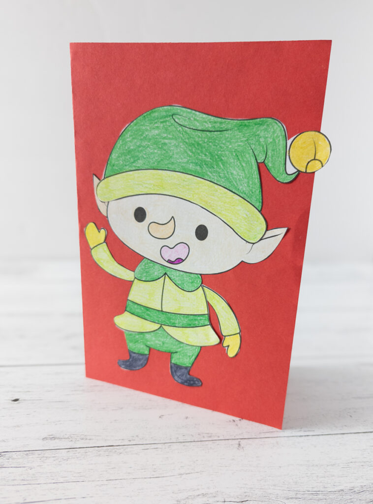 Cut and paste build an elf craft used to make a Christmas card. Elf is glued to front of red cardstock folded in half and standing up on table.