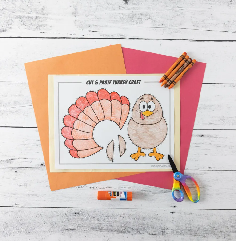 Build a turkey cut and paste craft colored in and laying on top of orange and red construction paper. Glue stick, scissors, and a few crayons are arranged around the paper.