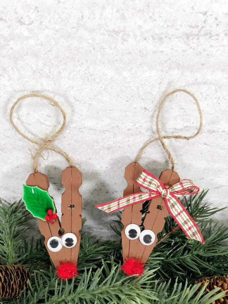 Two reindeer ornaments made out of clothespins leaning up against evergreen garland.