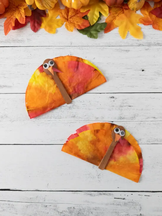 Two finished coffee filter turkey crafts made with mini spoons and dyed coffee filters. Fall leaves and pumpkins along the top.