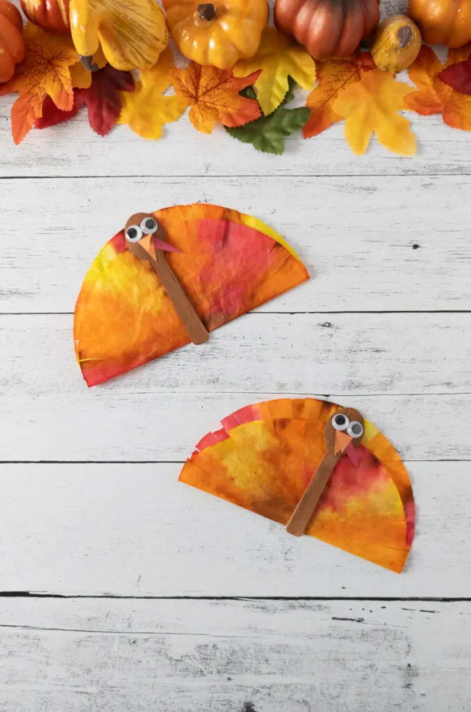 Two finished coffee filter turkey crafts made with mini spoons and dyed coffee filters. Fall leaves and pumpkins along the top.