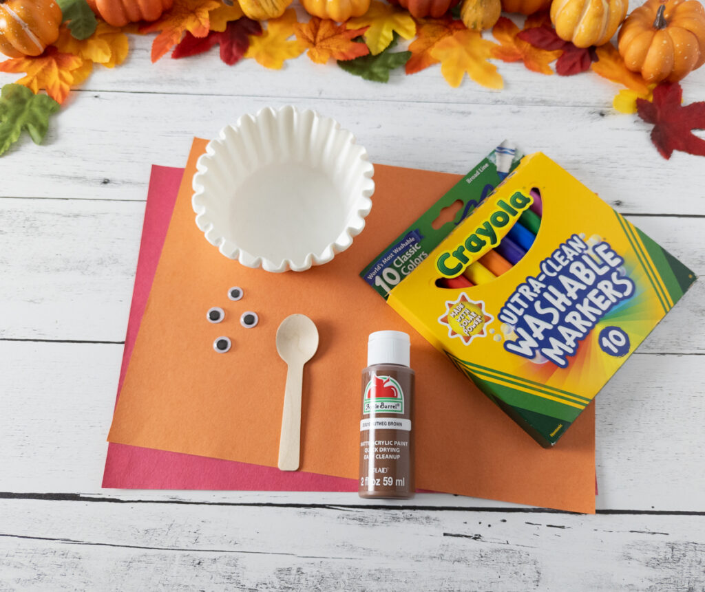 Supplies for turkey coffee filter craft includes red and orange paper, googly eyes, mini wooden spoon, brown paint, white coffee filters, and washable markers.
