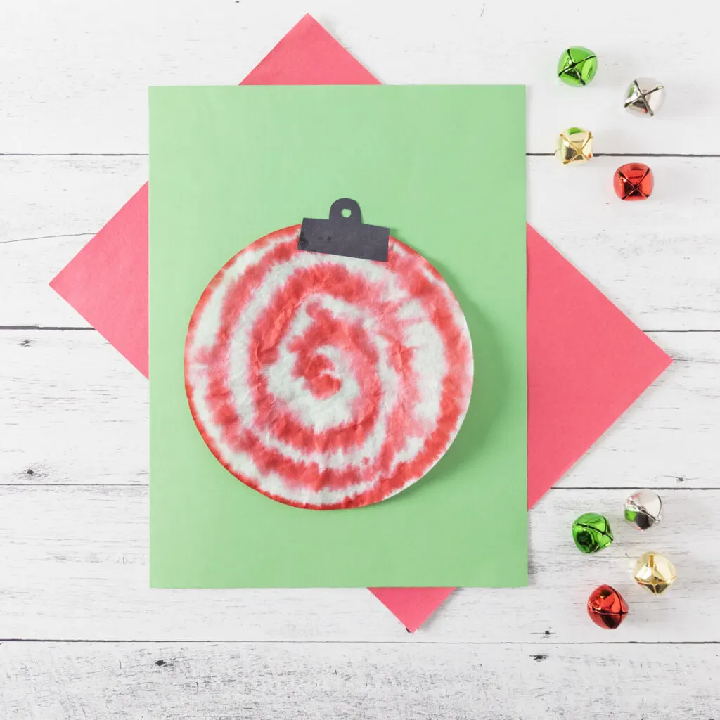 Coffee filter colored to look like a peppermint swirl ornament laying on green and red papers with jingle bells off to the side.