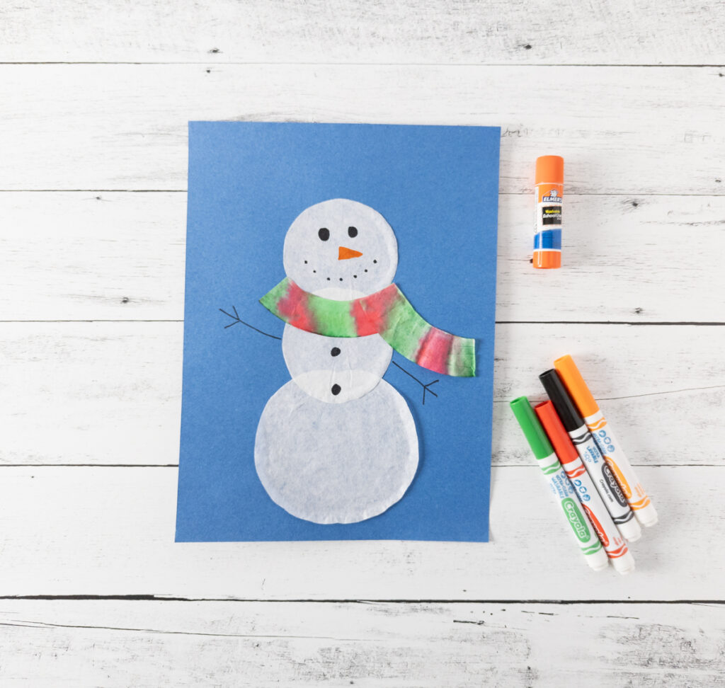 Finished coffee filter snowman with a red and green scarf on blue construction paper. Markers and glue stick laying next to it.