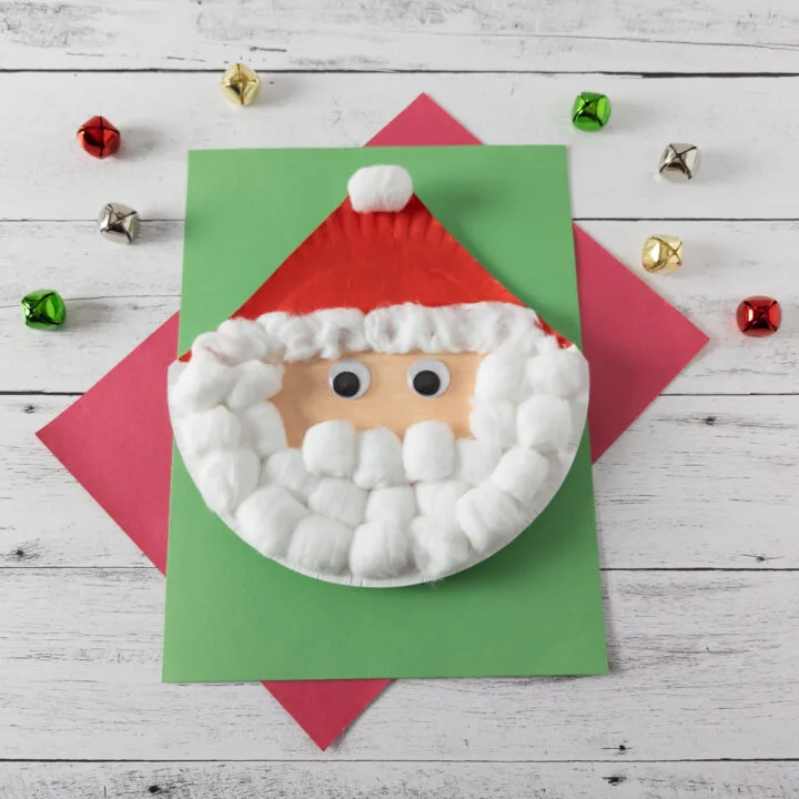 Paper plate Santa with cotton ball beard with top of plate cut to a point for his hat. Craft is laying on top of green and red paper with jingle bells scattered around.