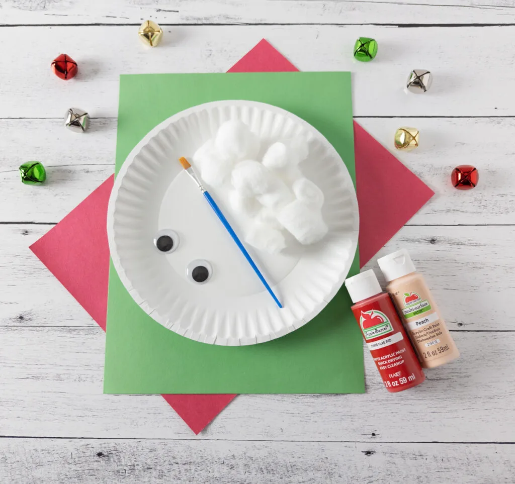 Supplies for Santa craft project laying on top of red and green paper. Paper plate, cotton balls, paint brush, googly eyes, red paint, and peach paint.