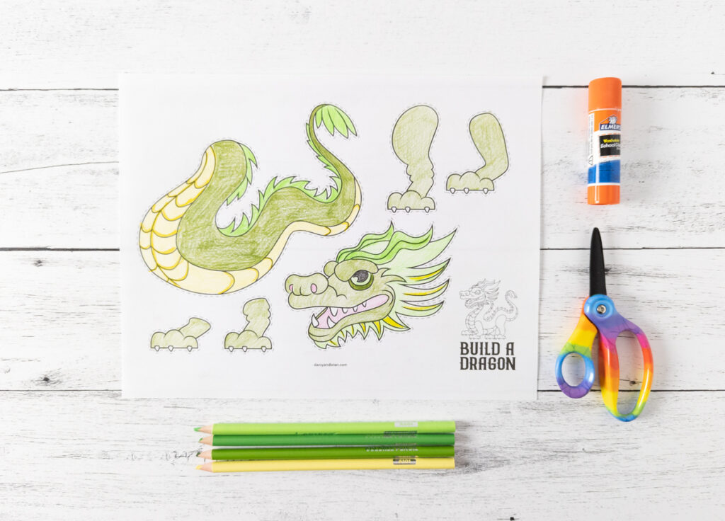 Printed out build a dragon is colored in with green and yellow. Colored pencils lay below paper. Scissors and glue stick lined up on the right.