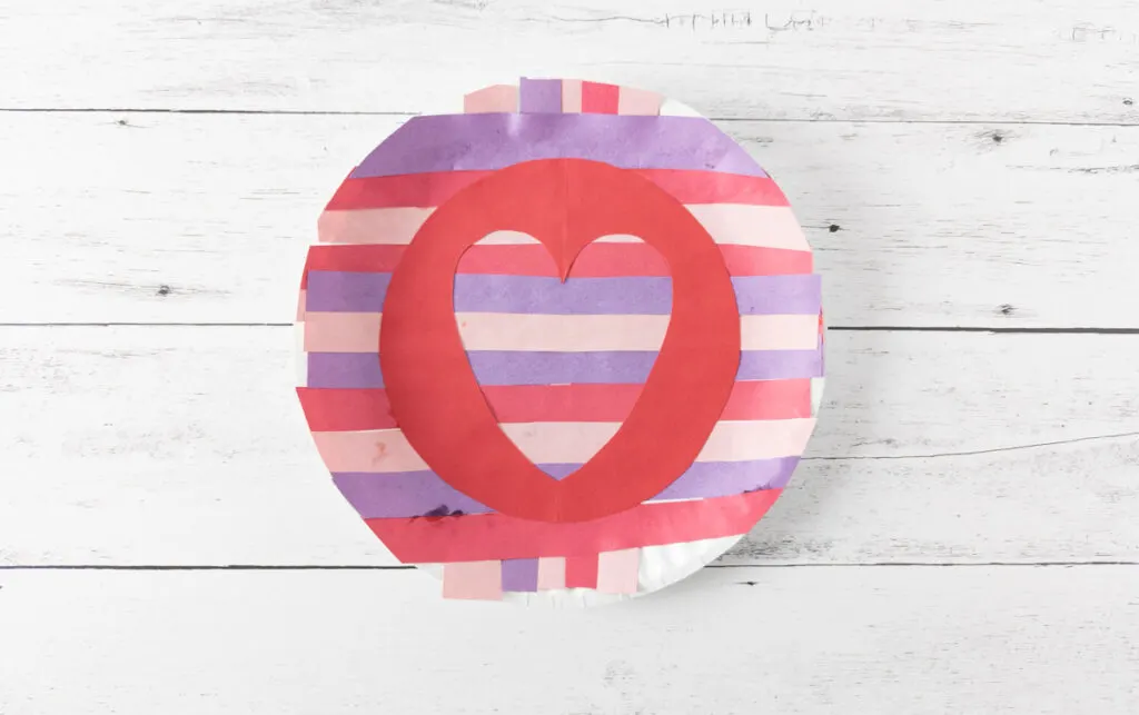 Large paper plate covered with red, purple, and pink strips of construction paper. On top of that is a red circle with a heart cut out framing the stripes in the heart.