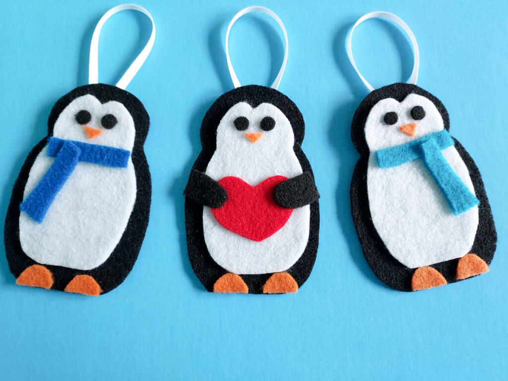 Three penguin kid crafts made with felt laying on blue paper. Two have scarves and one is holding a heart.