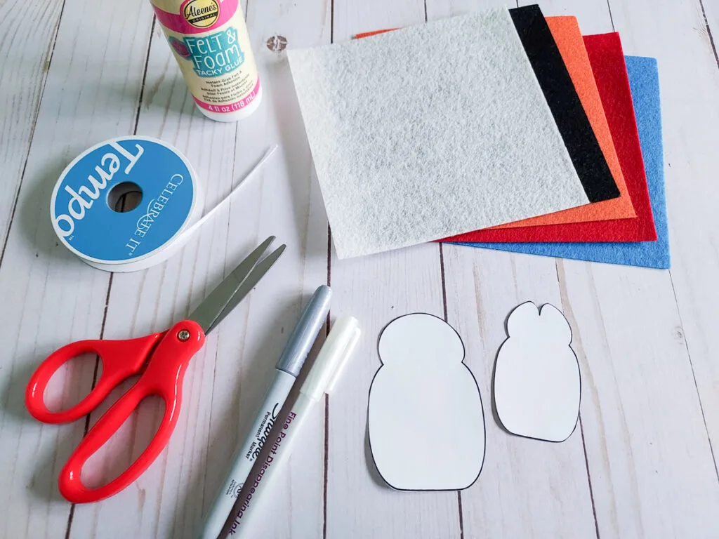 Felt penguin craft supplies on table. Includes felt squares in different colors, paper template, markers, scissors, tacky glue, and ribbon.