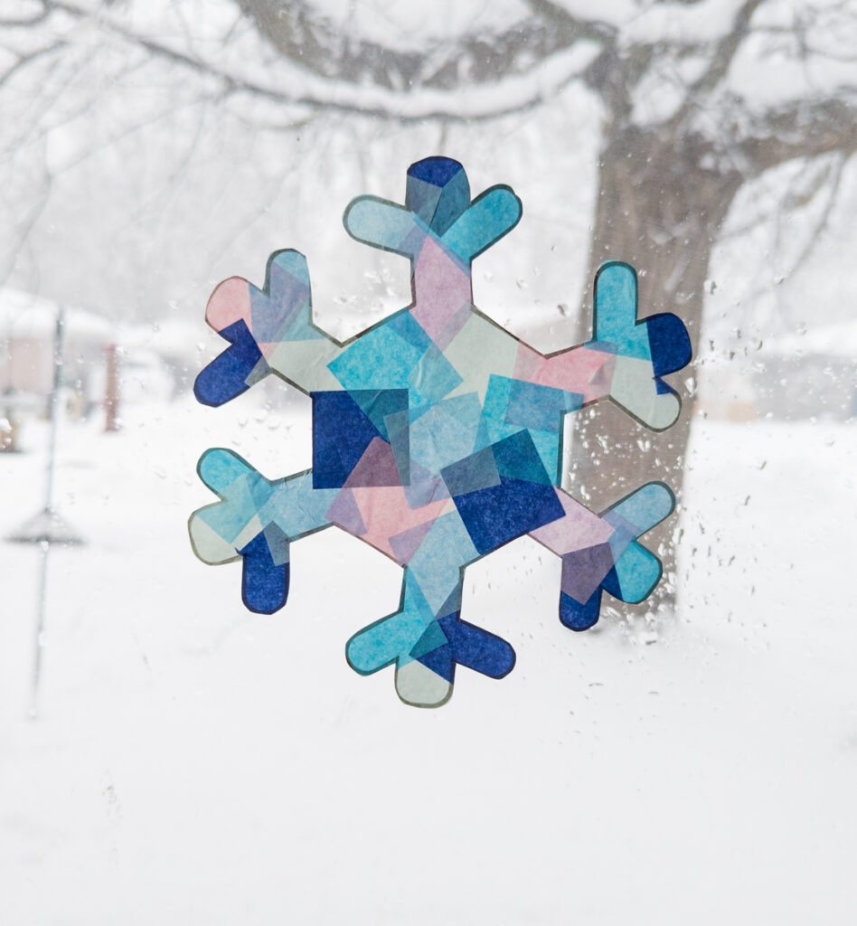 Close view of snowflake shaped suncatcher made with tissue paper in a snowy window.