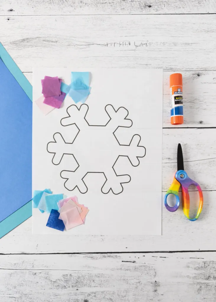 Snowflake craft template printed out with assorted small squares of tissue paper, glue stick, and scissors along it.