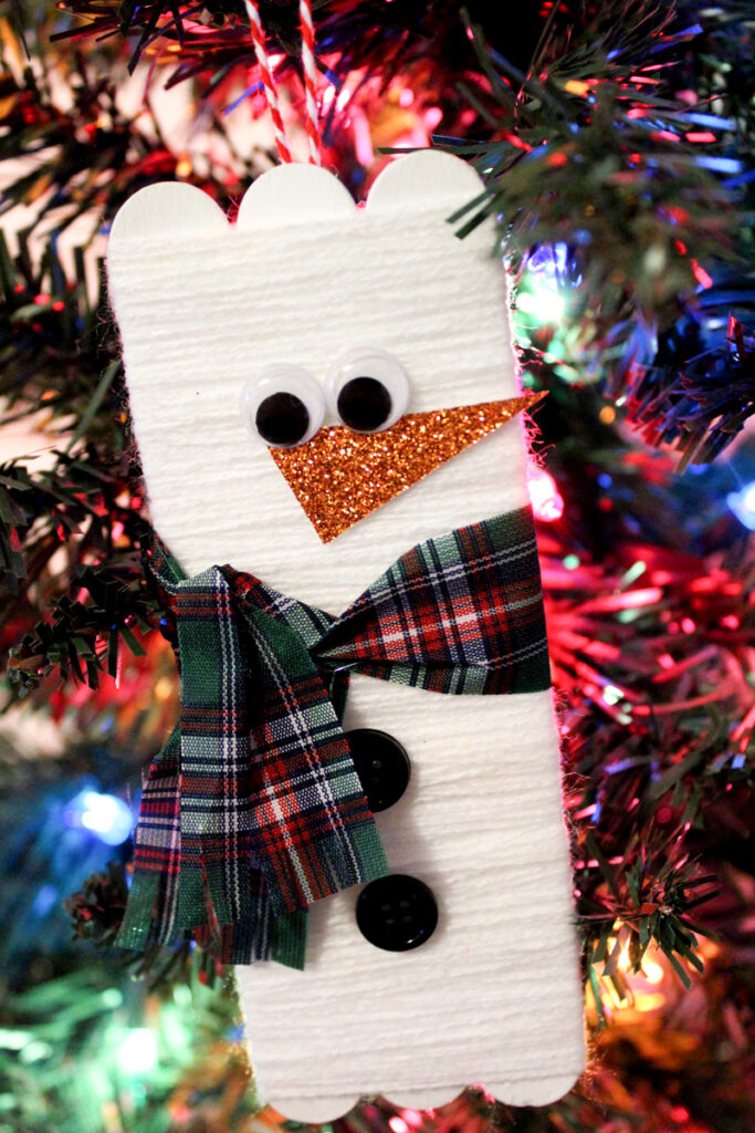 Completed snowman popsicle stick craft made into an ornament and hanging from a Christmas tree.