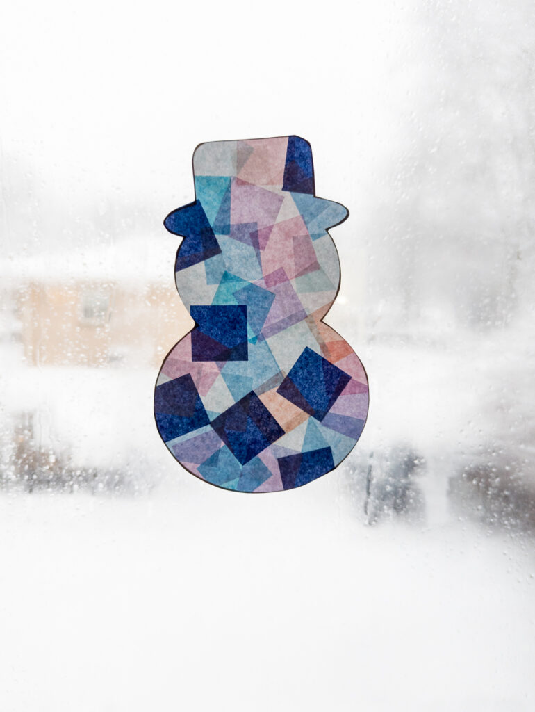 Finished tissue paper snowman suncatcher taped to window on a bright snowy day.