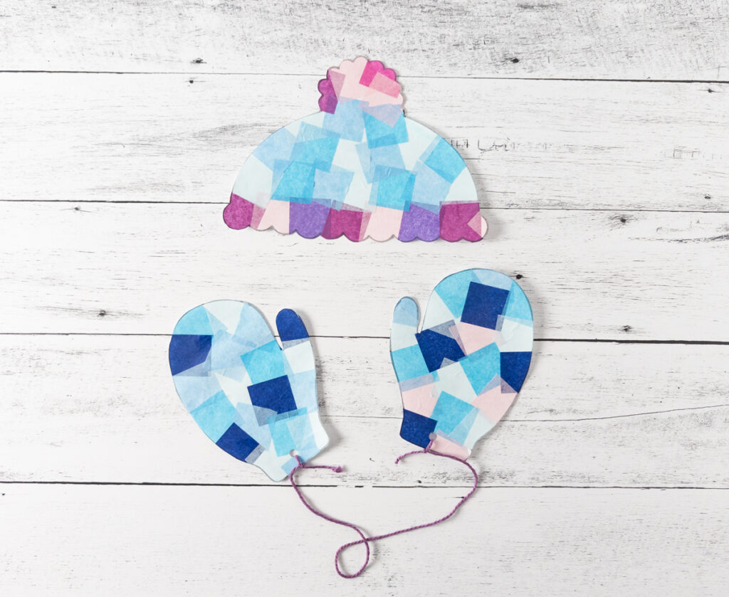 Winter hat and mittens suncatchers cut out. Mittens hole punched and tied together with purple yarn.