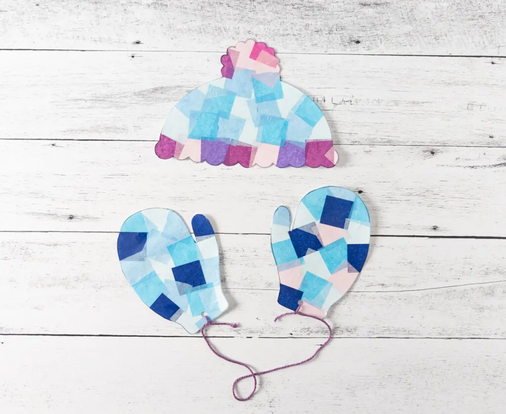 Winter hat and mittens suncatchers cut out. Mittens hole punched and tied together with purple yarn.