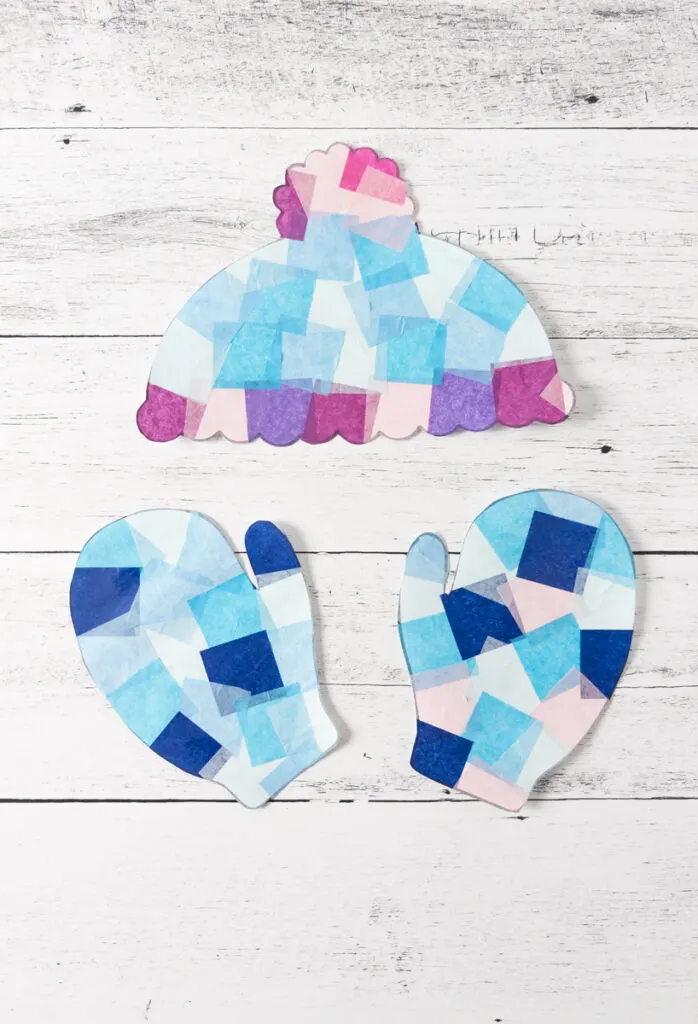 winter suncatcher crafts featuring a hat and mittens are covered in tissue paper and cut out laying on a table.
