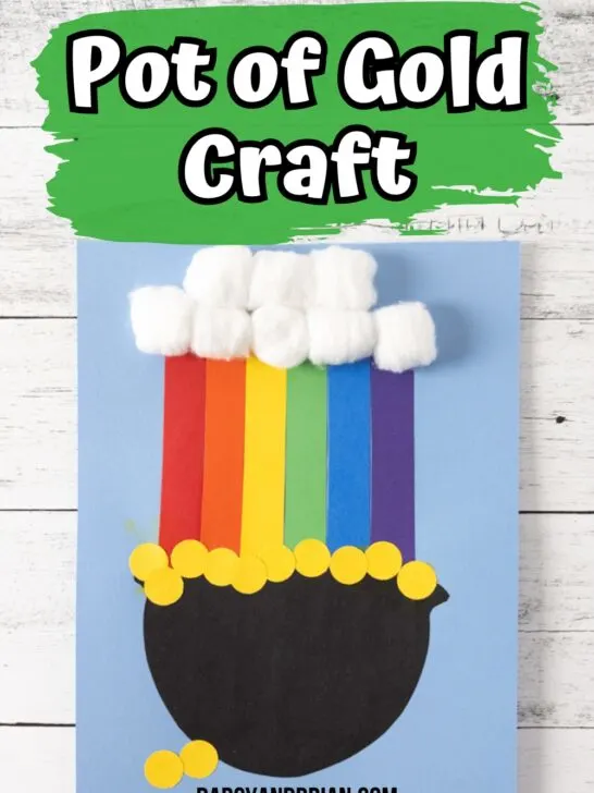 Pot of gold with a rainbow coming out of it made out of construction paper with cotton ball cloud. White text on green brush stroke says Pot of Gold Craft at the top. Project is glued onto a light blue piece of cardstock paper.