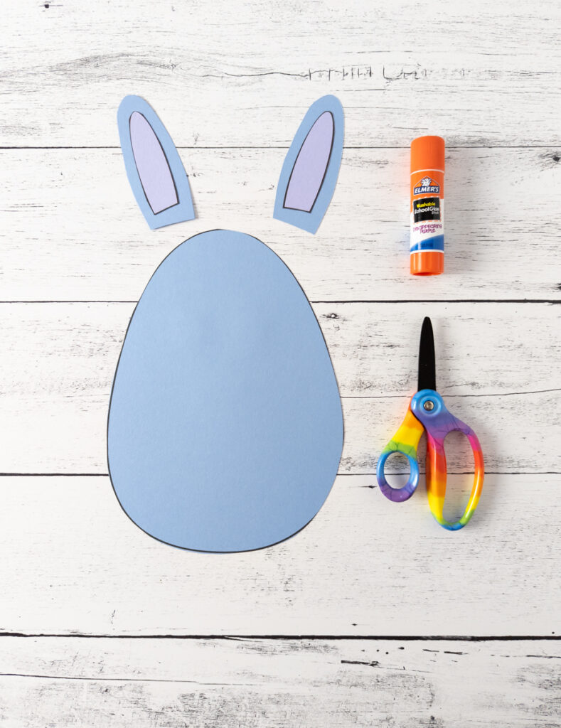 Egg shape cut out of blue paper. Light purple inner ear pieces glued onto blue ears. They are laying above the paper egg. Glue stick and scissors on the right side.