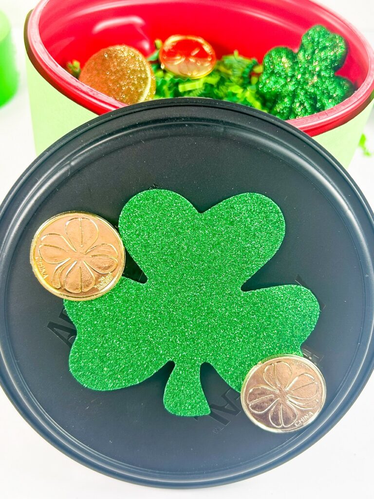 Black lid leaning against container and decorated with two gold coins and a large glittery shamrock.