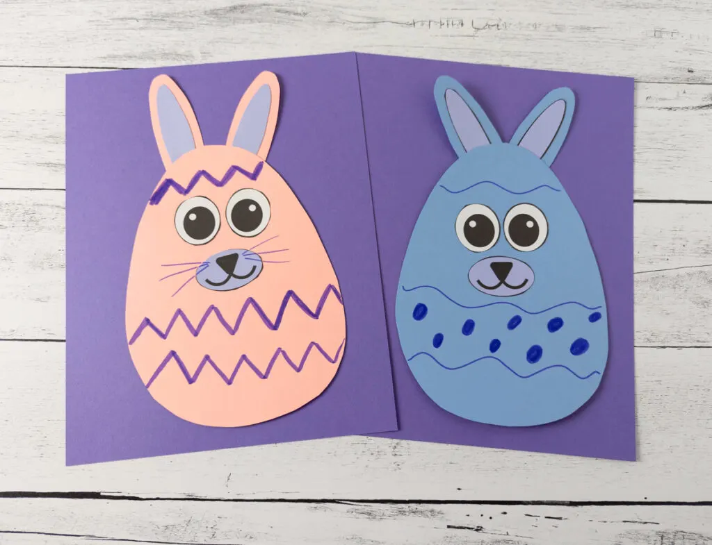 Two completed bunny egg craft projects made out of colored cardstock paper and decorated with markers. Pink and lavender bunny on purple paper. Blue and lavender bunny on purple paper.