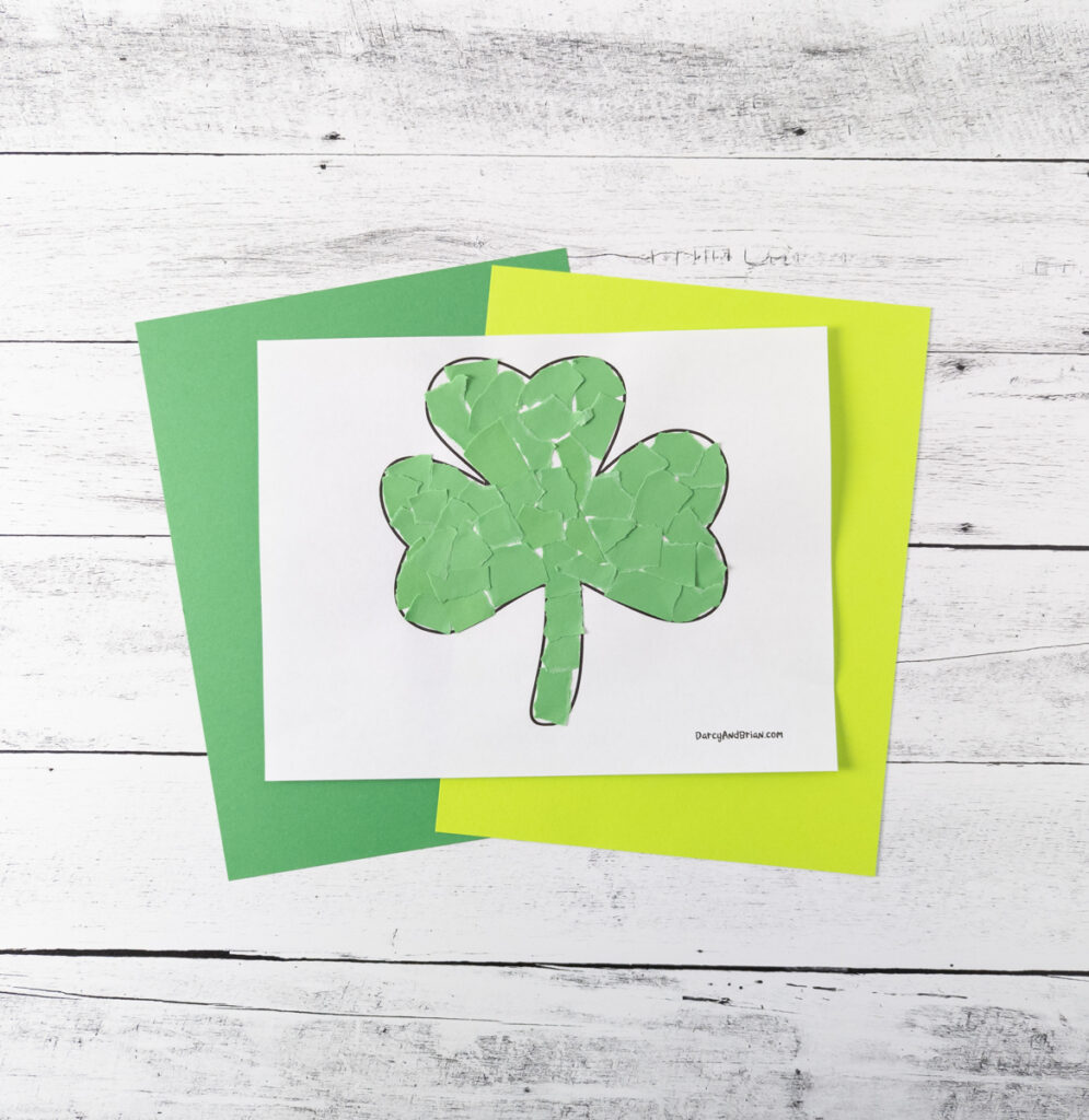 Shamrock template filled with ripped up pieces of green paper. Craft template is laying on top of green and yellow papers.