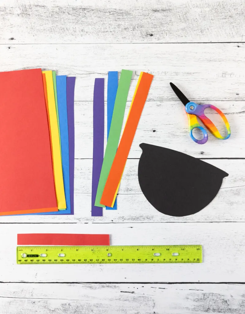 Construction paper in rainbow colors cut into one inch wide strips and laying next to pot cut out of black paper. Red piece of paper laying along ruler showing it was trimmed down to be six inches long.
