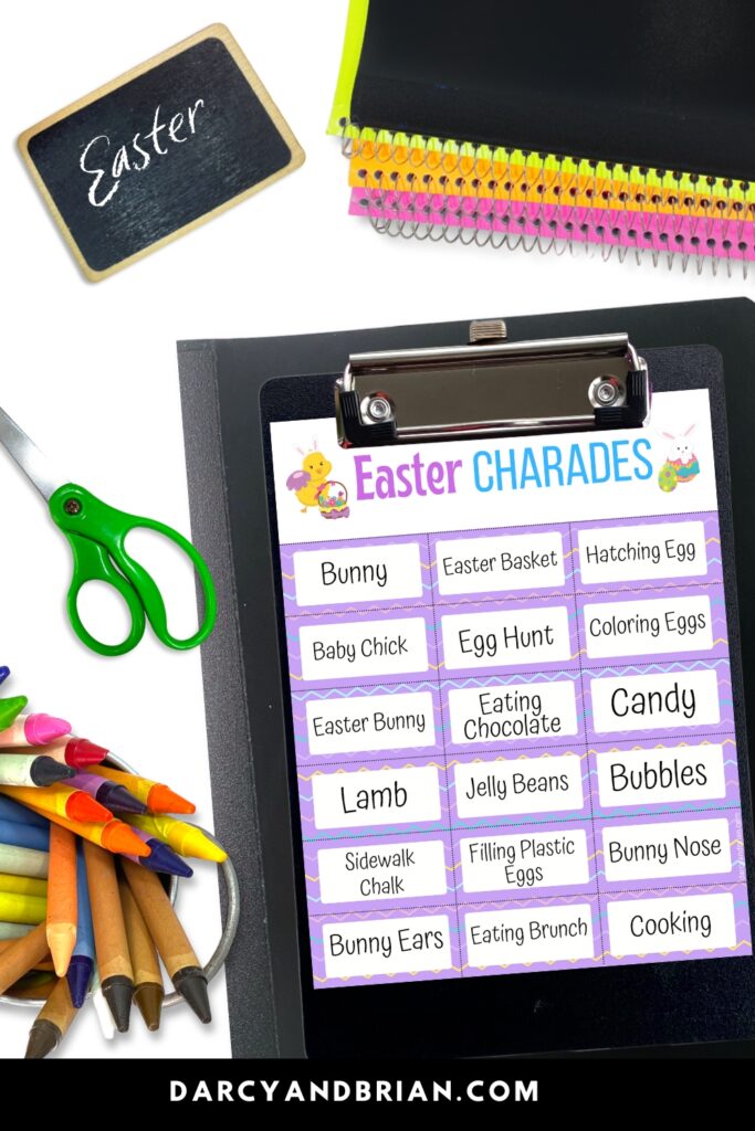 One page of Easter charades prompts on a black clipboard. A stack of colorful notebooks at the top next to a small chalkboard that says Easter. A pair of scissors and a bunch of crayons also next to it.