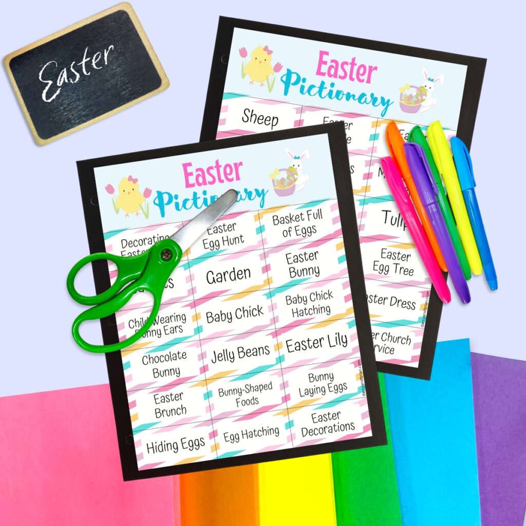 Mockup of printable Easter themed Pictionary game words laying on top of colorful construction paper. Pair of green handled scissors and markers laying over the pages. A small chalkboard with the word Easter is in the upper left corner.