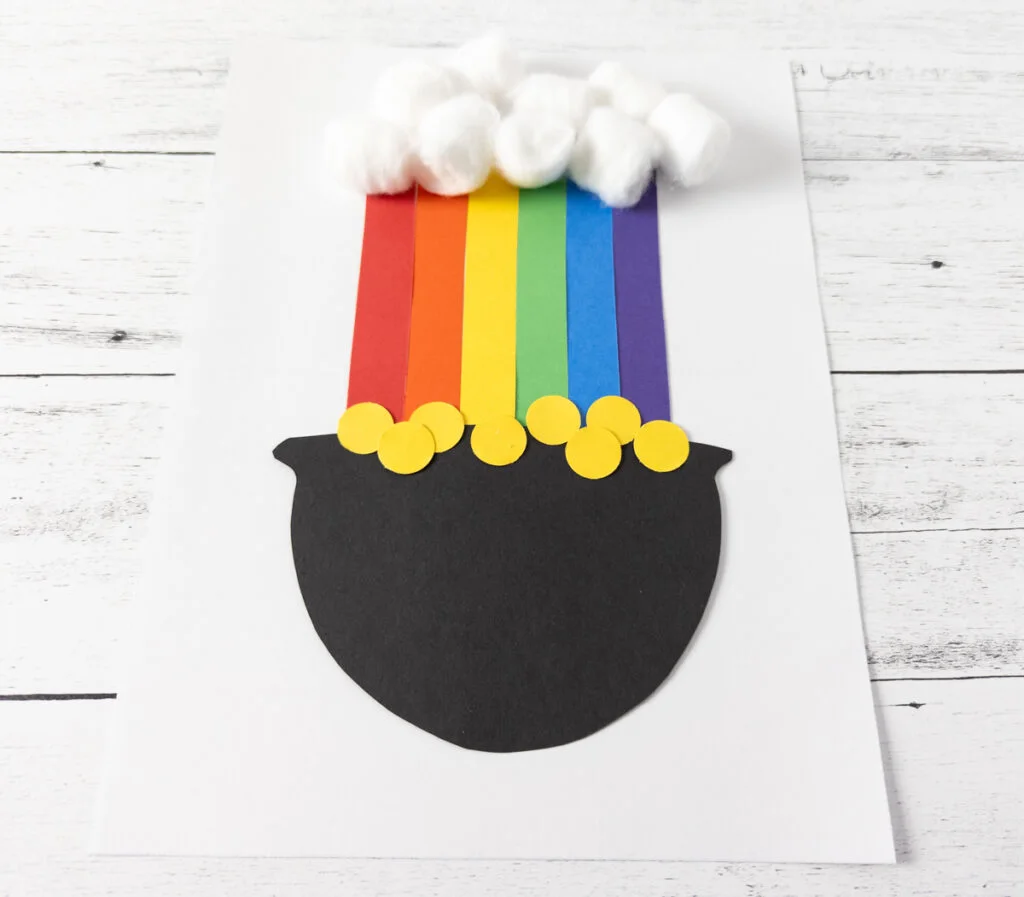 Tilted down angle view of rainbow and pot of gold made out of paper and cotton balls glued to white paper.