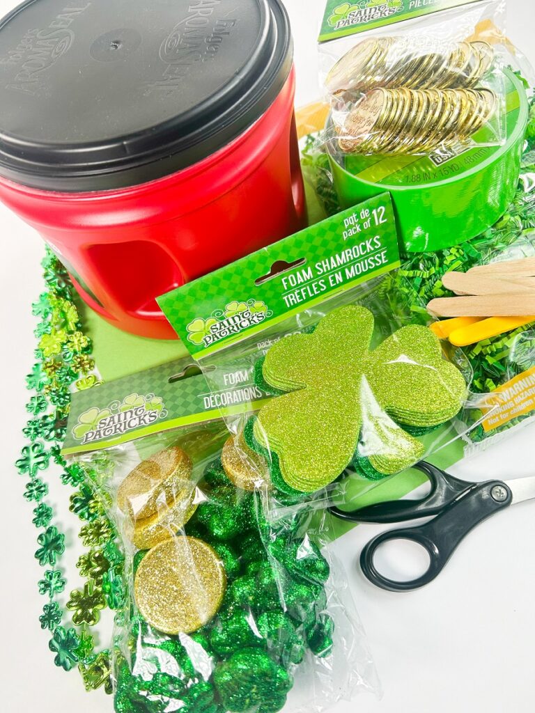 Supplies to make a trap for Saint Patrick's Day including a plastic coffee container with lid, shamrock cutouts, shamrock beads, coins, green paper shred.