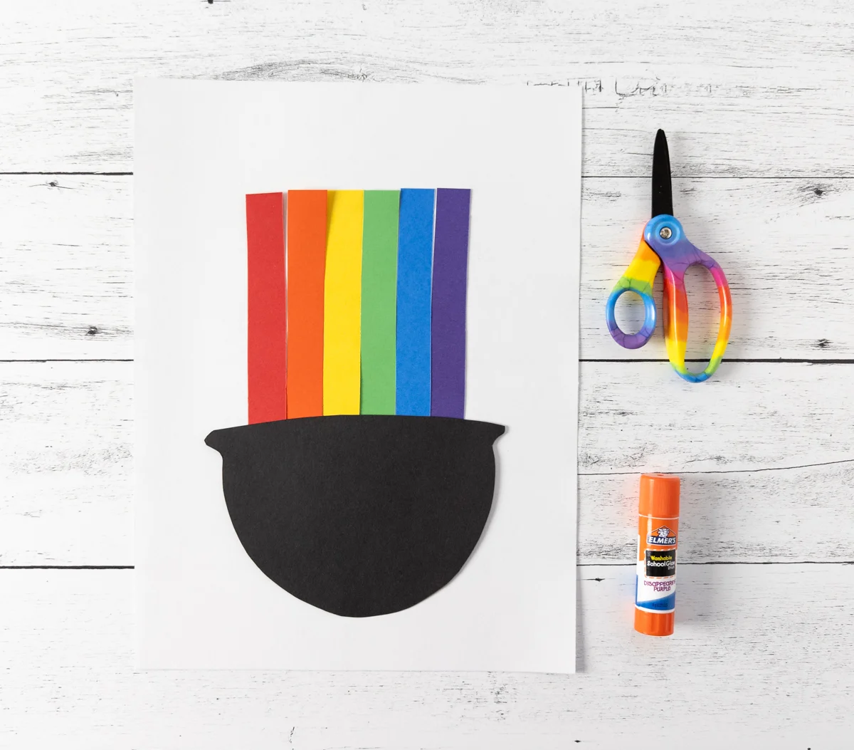 Black paper cauldron shape cut out and glued down overlapping the bottom of vertical rainbow stripes.