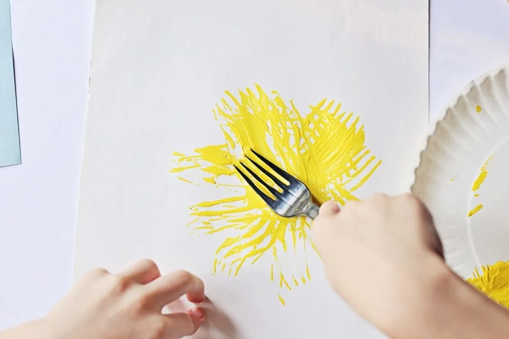 White child's hand using a fork to apply yellow paint to white paper.