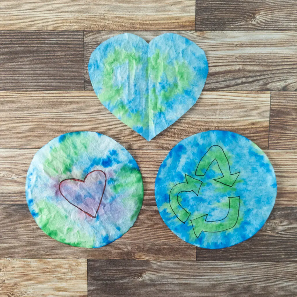 Three versions of Earth Day coffee filter art. One is blue and green cut into a heart. One is the Earth with a red heart in the center. One is blue with a green recycle symbol in the center.