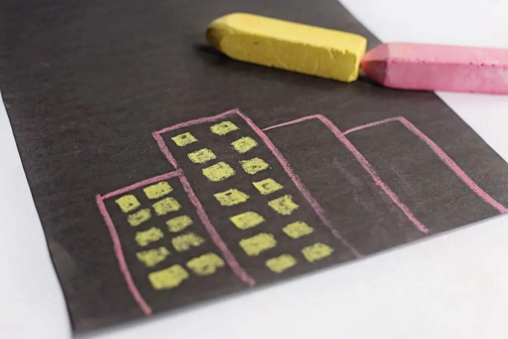Four rectangles drawn on black paper using pink chalk. Two buildings have yellow square windows drawn to look like tall business buildings.