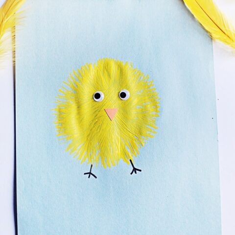 Top down view of finished fork painted chick. Yellow baby chick on light blue paper with yellow feathers around it.