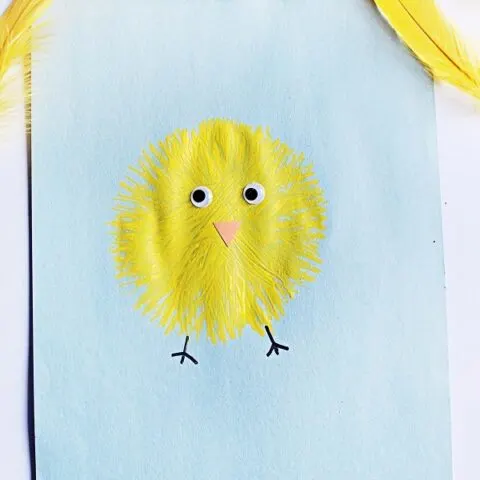 Top down view of finished fork painted chick. Yellow baby chick on light blue paper with yellow feathers around it.