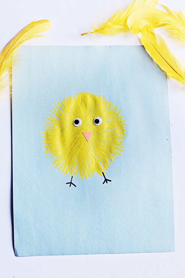 Top down view of finished fork painted chick. Yellow baby chick on light blue paper with yellow feathers around it.