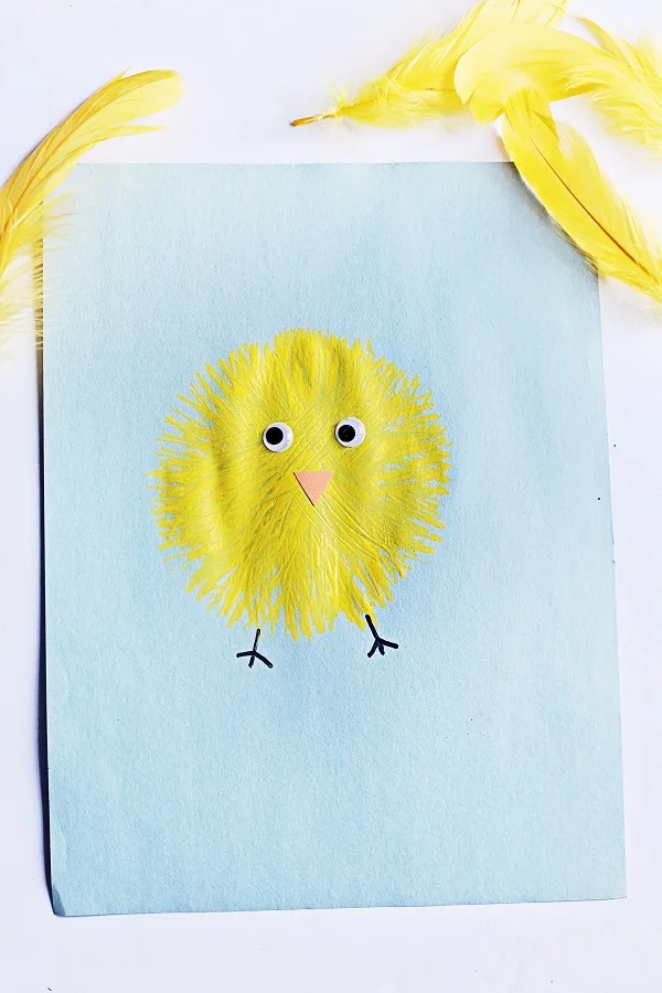 Top down view of finished fork painted chick. Yellow baby chick on light blue paper with yellow feathers around it.