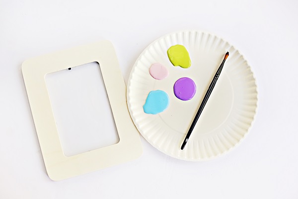Plain wooden craft picture frame laying next to paper plate with four different colors of paint on it and a paint brush.