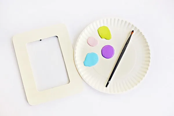 Plain wooden craft picture frame laying next to paper plate with four different colors of paint on it and a paint brush.