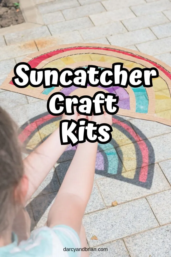 Child holding out a rainbow suncatcher made with cardboard and clear plastic making a colorful rainbow on the sidewalk. White text in the center says Suncatcher Craft Kits