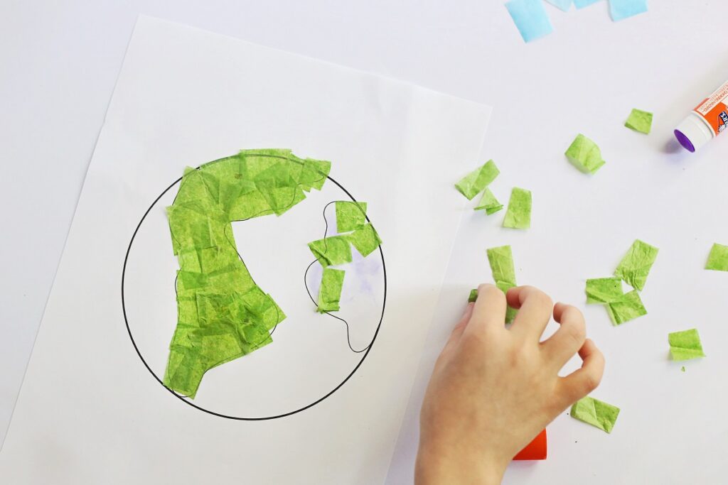 White child's hand carefully gluing green tissue paper pieces to Earth craft template.
