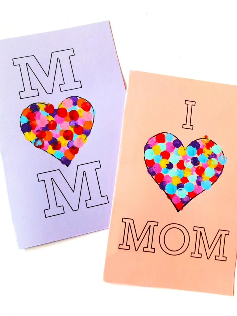 Two finished Mother's Day heart cards decorated with colorful fingerprints. One card on lavender paper says Mom with heart in the center. Pink card says I (heart) Mom.