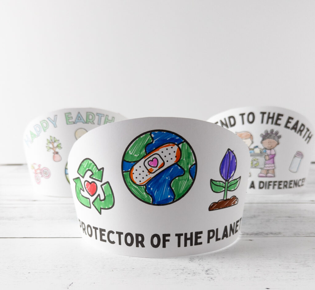 Three finished Earth Day crown headbands for kids arranged on a table. The one in front is colored with markers and says Protector of the Planet.